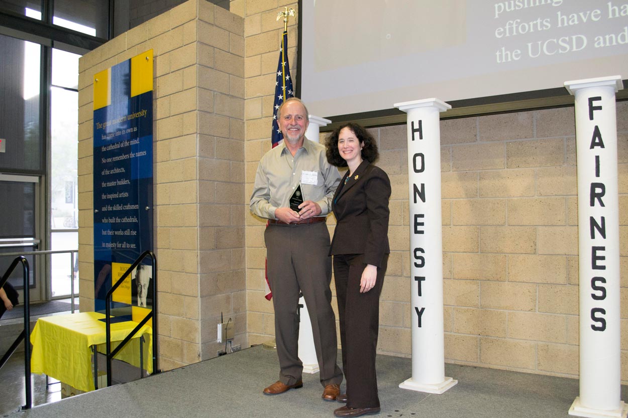 Jim Posakony holding his 2018 Integrity Award and standing next to UC San Diego Executive Vice Chancellor Elizabeth Simmons onstage.