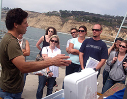 A group of graduate students standing on a boat in front of a male speaker