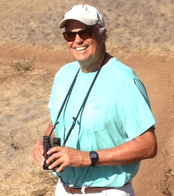 Ted Case in a blue shirt, holding binoculars standing in a field.